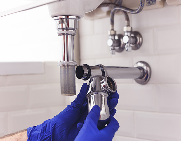 hands wearing blue gloves working on plumbing under a sink holding a shiny chrome pipe for a 22 millimeter connection and drainage system