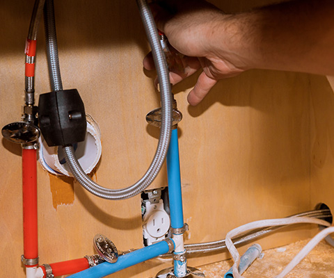 hand reaching for plumbing connections under sink with red and blue pipes showing installation process for water lines 2 plumbing tasks needed